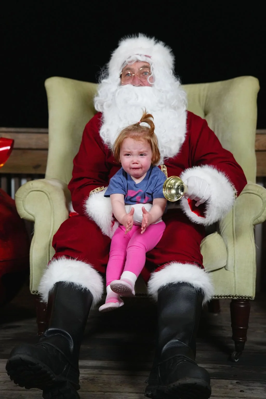 A young girl with red hair and pink pants sitting on Santa Claus's lap, who is dressed in traditional red and white, holding a golden bell, inside a room with a wooden floor and dark background.