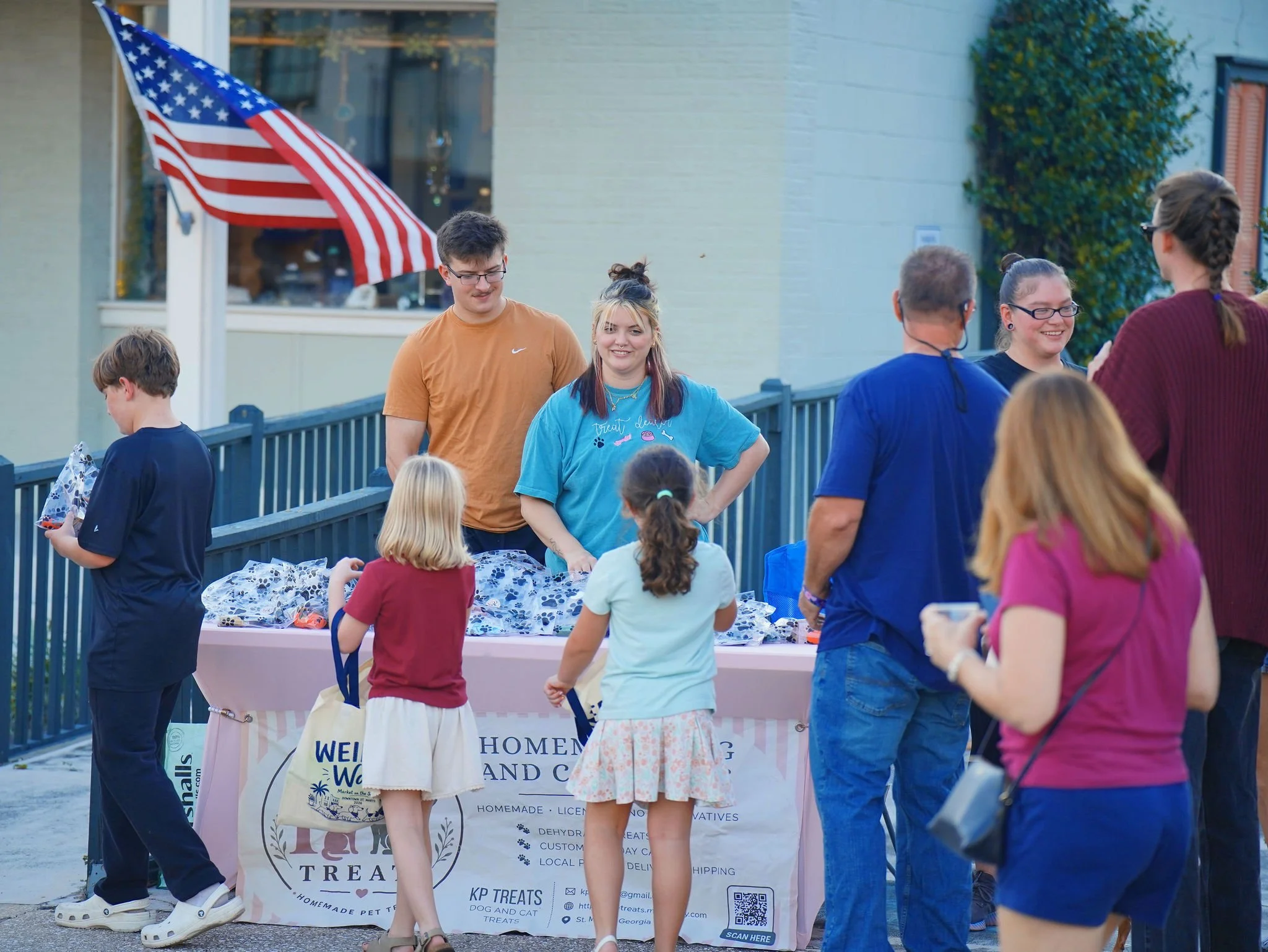 A group of people, including children and adults, gather outdoors around a table at a community event. An American flag is visible in the background, waving near a tan building. The table has a pink tablecloth with a sign advertising homemade pet tre