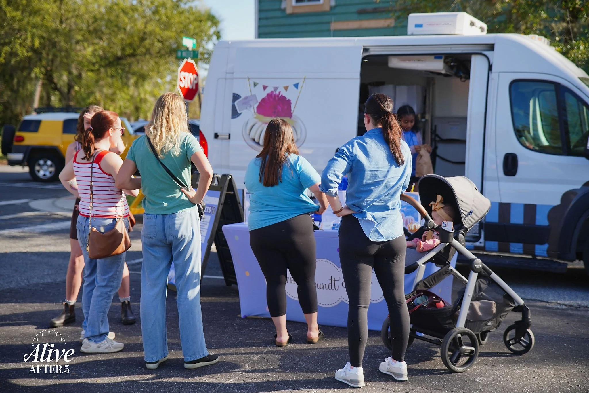 A group of women and a baby in a stroller line up at a mobile ice cream truck on a sunny day, with trees and parked cars in the background.