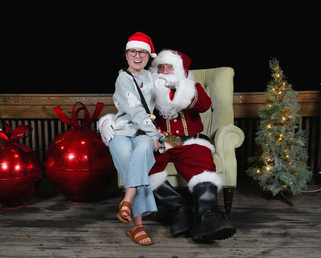A person dressed as Santa Claus sitting on a chair with a woman wearing a Santa hat, both smiling, in front of a Christmas tree and large red gift boxes, on a wooden deck with a black background.