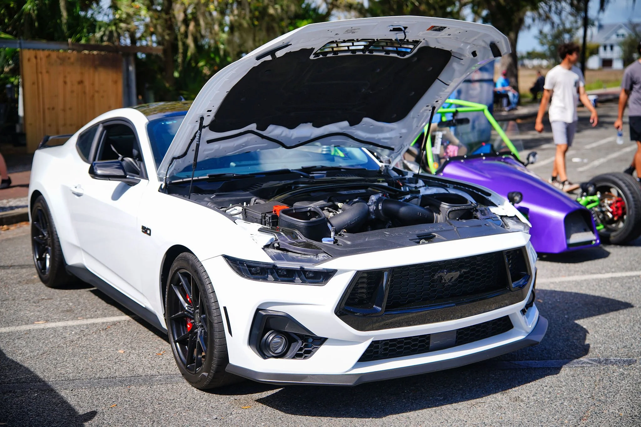 A white Ford Mustang with its hood open, displaying the engine at an outdoor car event, with other vehicles and people in the background.