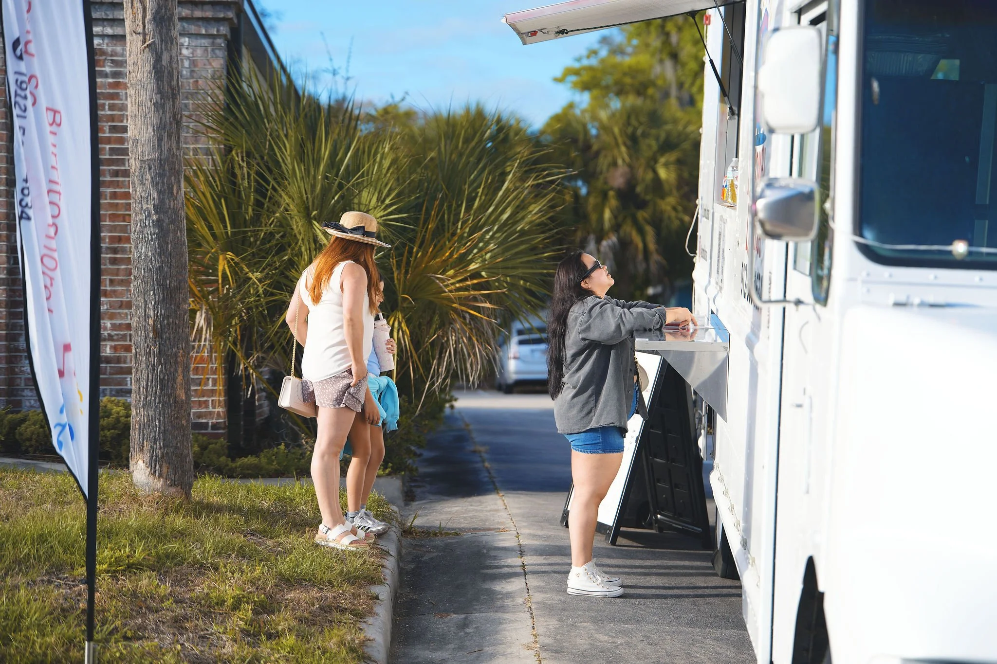 Three women are standing in line at a food truck, with palm trees and parked cars in the background.