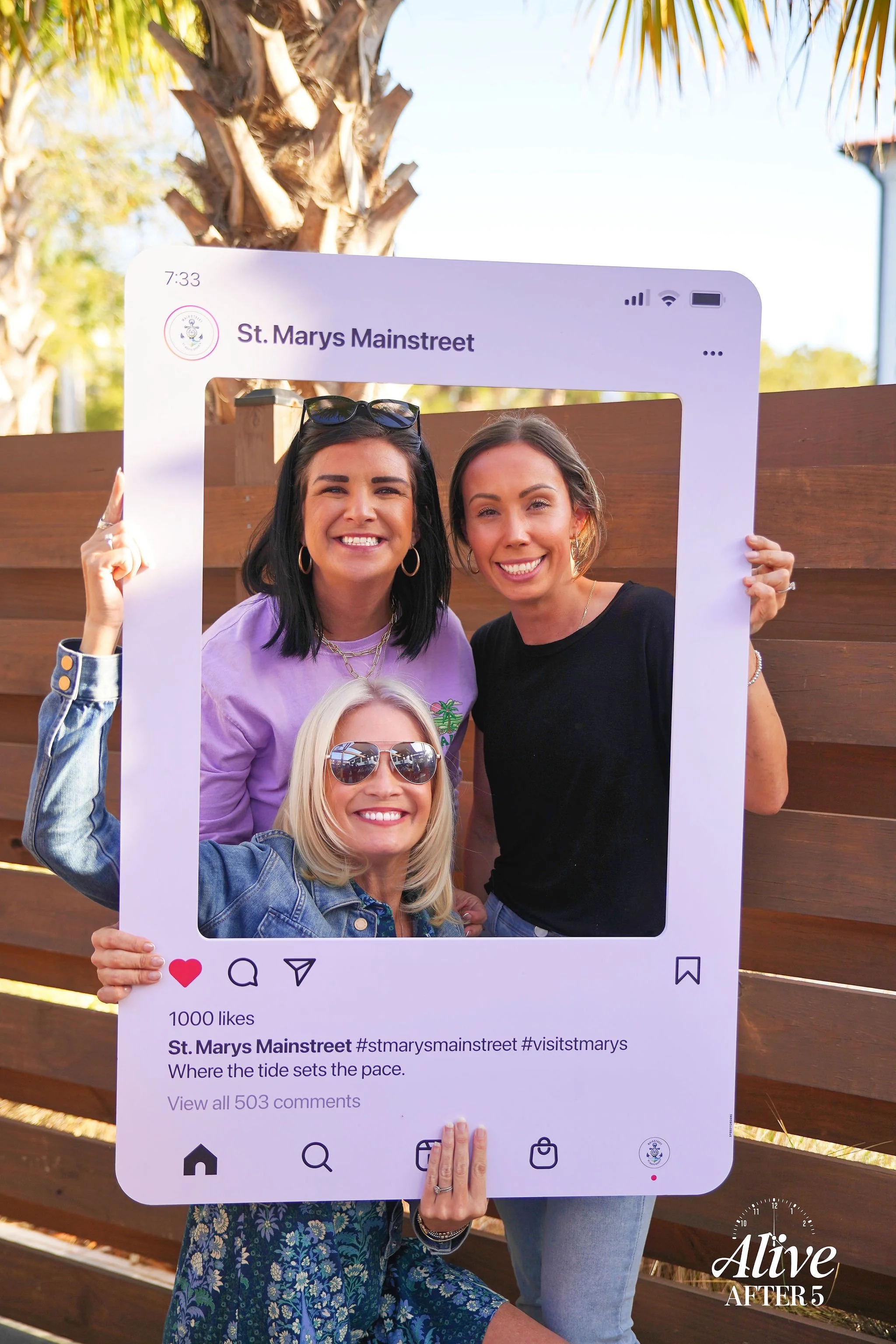 Three women smiling and posing behind a large Instagram photo frame prop at St. Marys Mainstreet outdoors with palm trees and a wooden fence in the background.