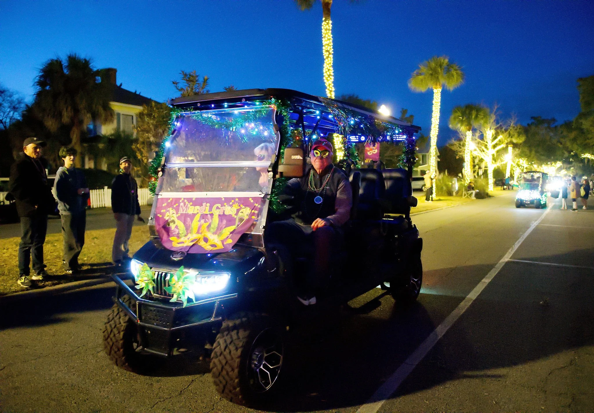 A decorated golf cart in a parade with a person wearing a Mardi Gras mask sitting inside. The golf cart is adorned with colorful lights, beads, and a Mardi Gras banner. Palm trees and streetlights illuminate the evening scene, with spectators standin