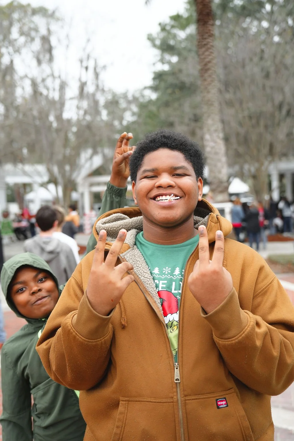 Two young boys outdoors, smiling at the camera. The boy in the foreground is making peace signs with both hands, wearing a brown jacket and green shirt. The boy in the background has a dark green hooded jacket, standing behind the first boy. Trees an