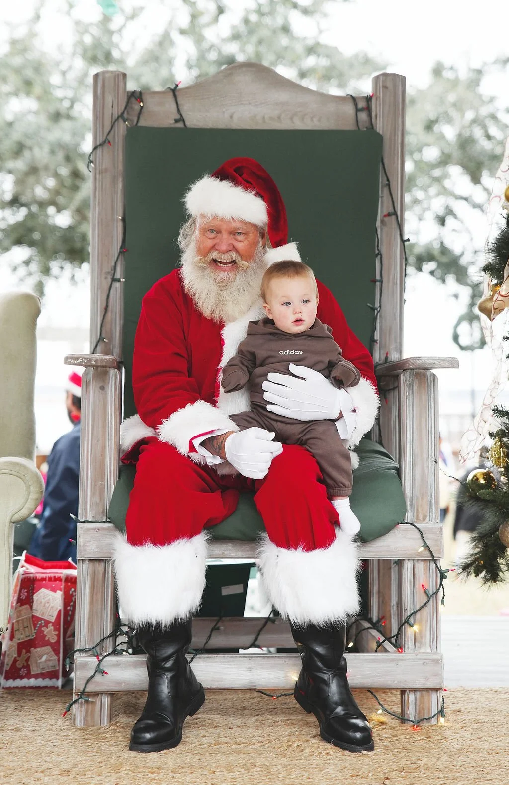 Santa Claus sitting on a large wooden chair with a green backrest, holding a young boy in his lap during a Christmas event. Santa is wearing a red suit with white fur trim, a red hat, and black boots. The boy is dressed in a brown Adidas outfit. The 
