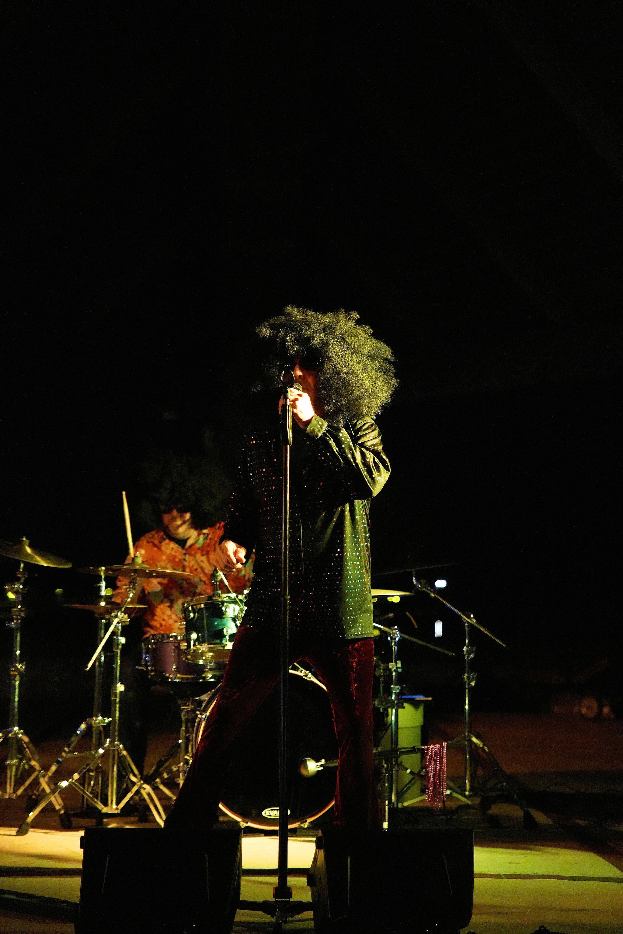 A performer with a large afro hairstyle singing into a microphone, wearing sunglasses and a shiny black shirt, with a drummer in the background during a nighttime outdoor music performance.