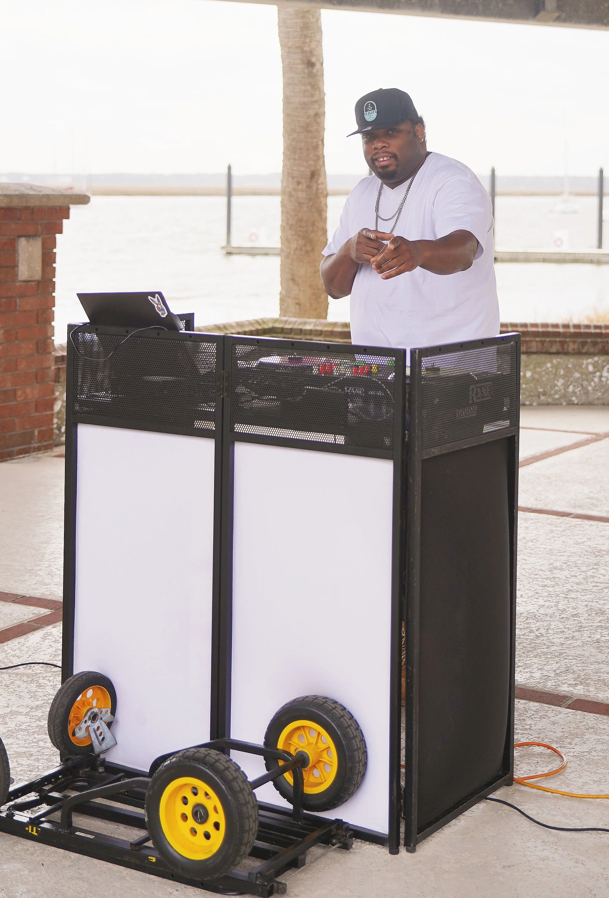 A DJ is standing behind a DJ booth with a laptop, wearing a cap and white t-shirt, outdoors near water with boats in the background.