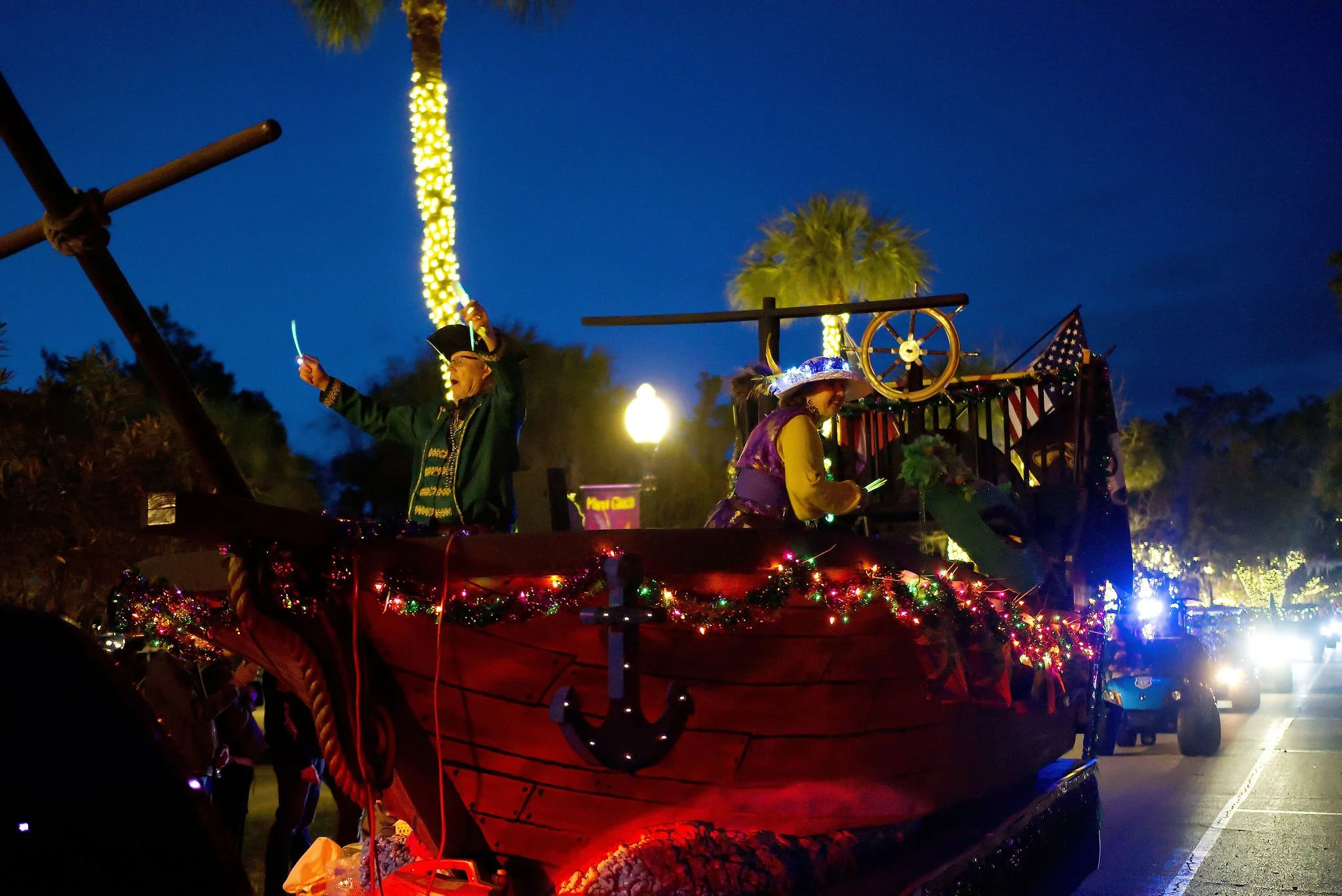 A decorated parade float with two people dressed in festive costumes, one of whom is wearing pirate attire, during a nighttime parade with palm trees and streetlights in the background.