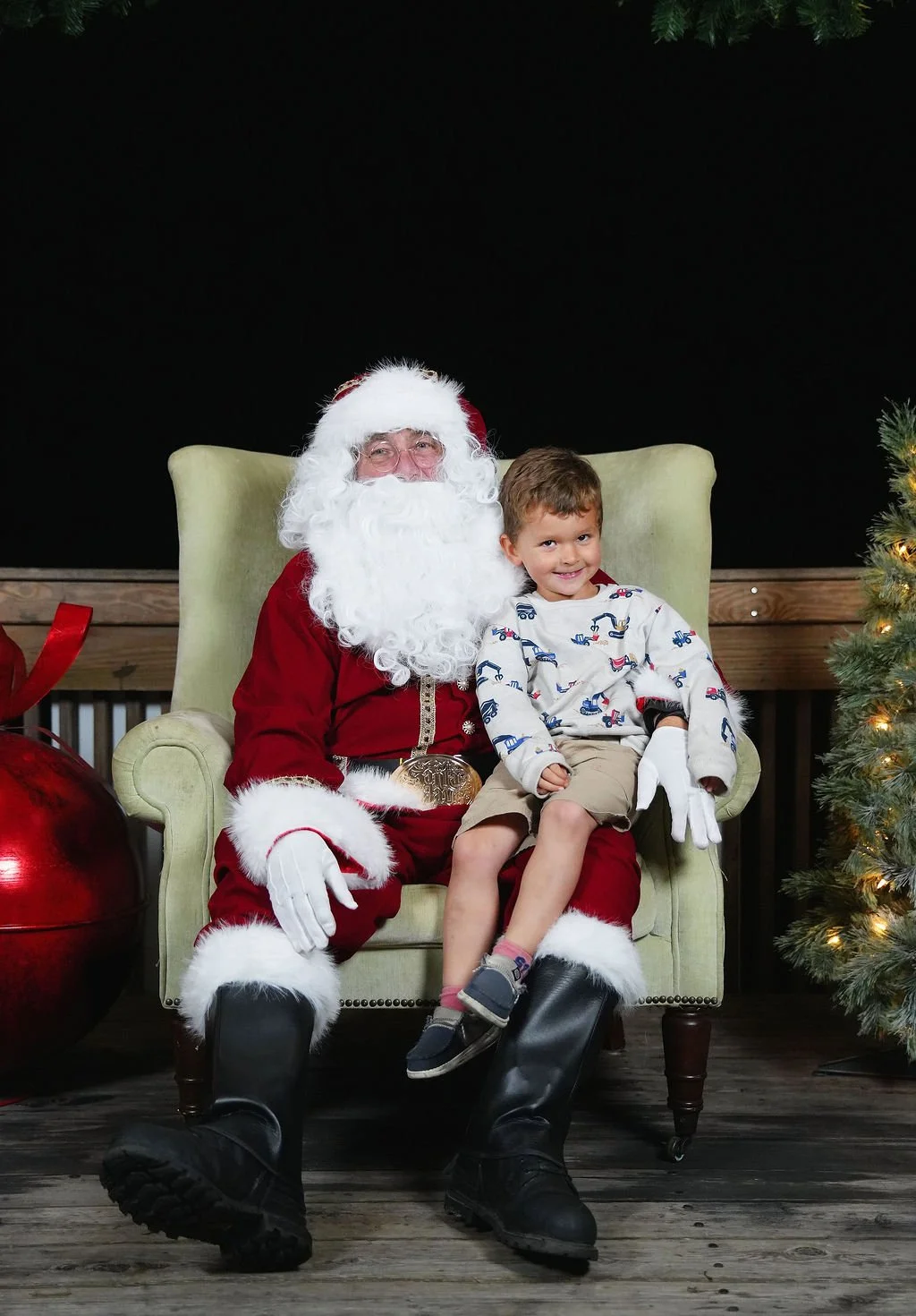 A young boy sitting on Santa Claus's lap, smiling, during a Christmas event. Santa is dressed in a traditional red suit with white fur trim, sitting in a green armchair, with Christmas decorations around, including a Christmas tree on the right and a