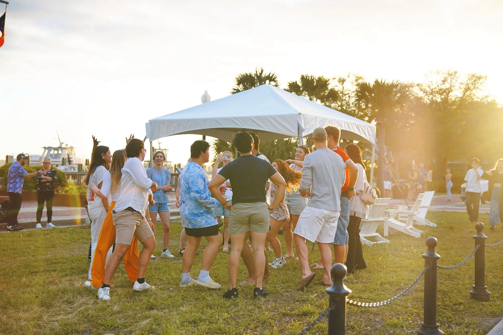 Group of people dancing and socializing on a grassy area at sunset, with a white canopy tent and benches nearby.