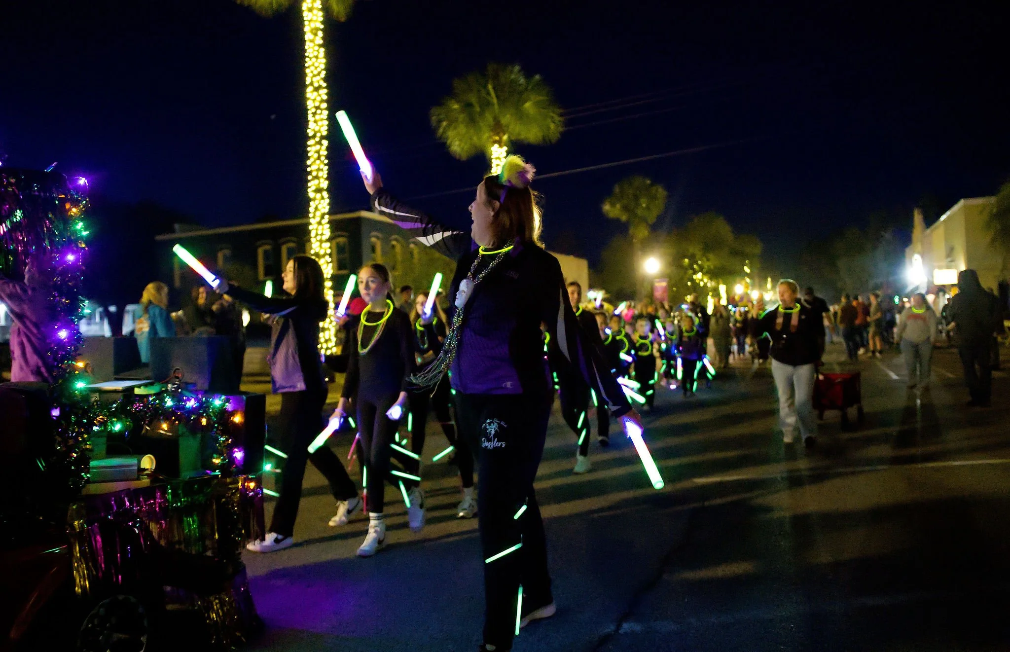 Nighttime parade with people in black clothing decorated with glow sticks, walking down a street with palm trees and buildings, illuminated by streetlights and decorated with colorful lights.