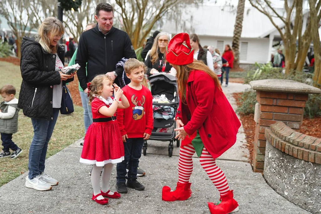 A woman dressed as an elf in red and white striped leggings and red elf shoes is entertaining children and adults at a holiday event outdoors, with children dressed in Christmas-themed clothing.