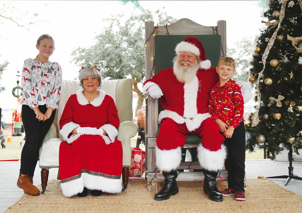 A family with children taking a photo with Santa Claus and Mrs. Claus at a Christmas event outdoors during daytime. Santa is seated in a chair, Mrs. Claus is seated nearby, and two children are standing on either side, all smiling.