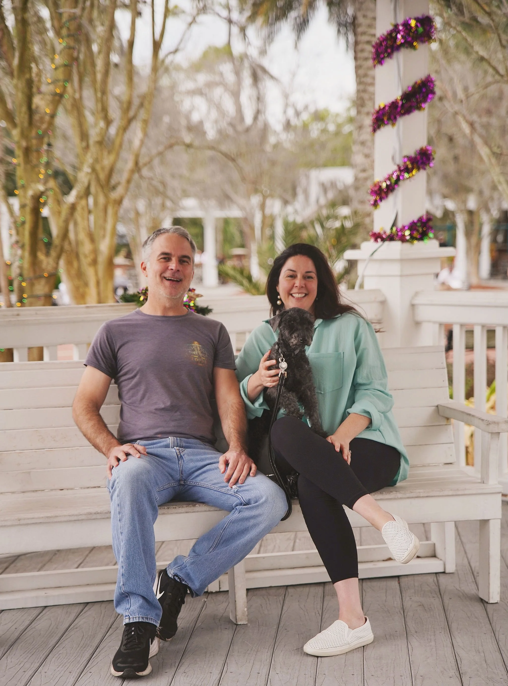 A smiling couple sitting on a white wooden bench outdoors with their dog. The man is wearing a gray t-shirt and jeans, and the woman is wearing a light green shirt and black leggings. The woman is holding a black dog. There are decorated trees and a 