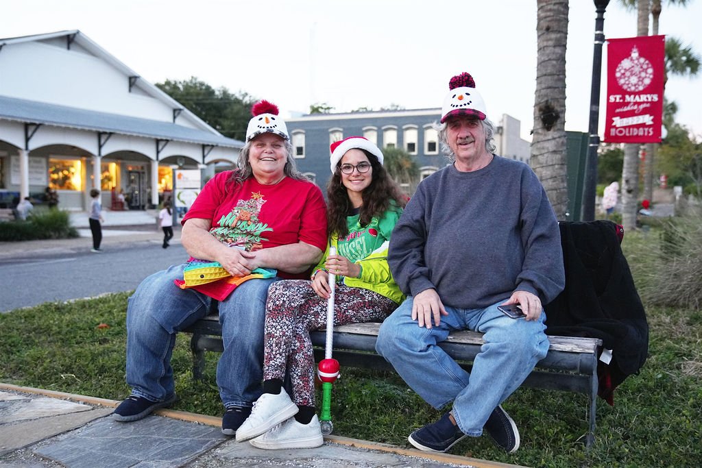 Three people sitting on a park bench wearing Christmas hats with snowman designs, celebrating the holidays in a small town with Christmas decorations and festive clothing.