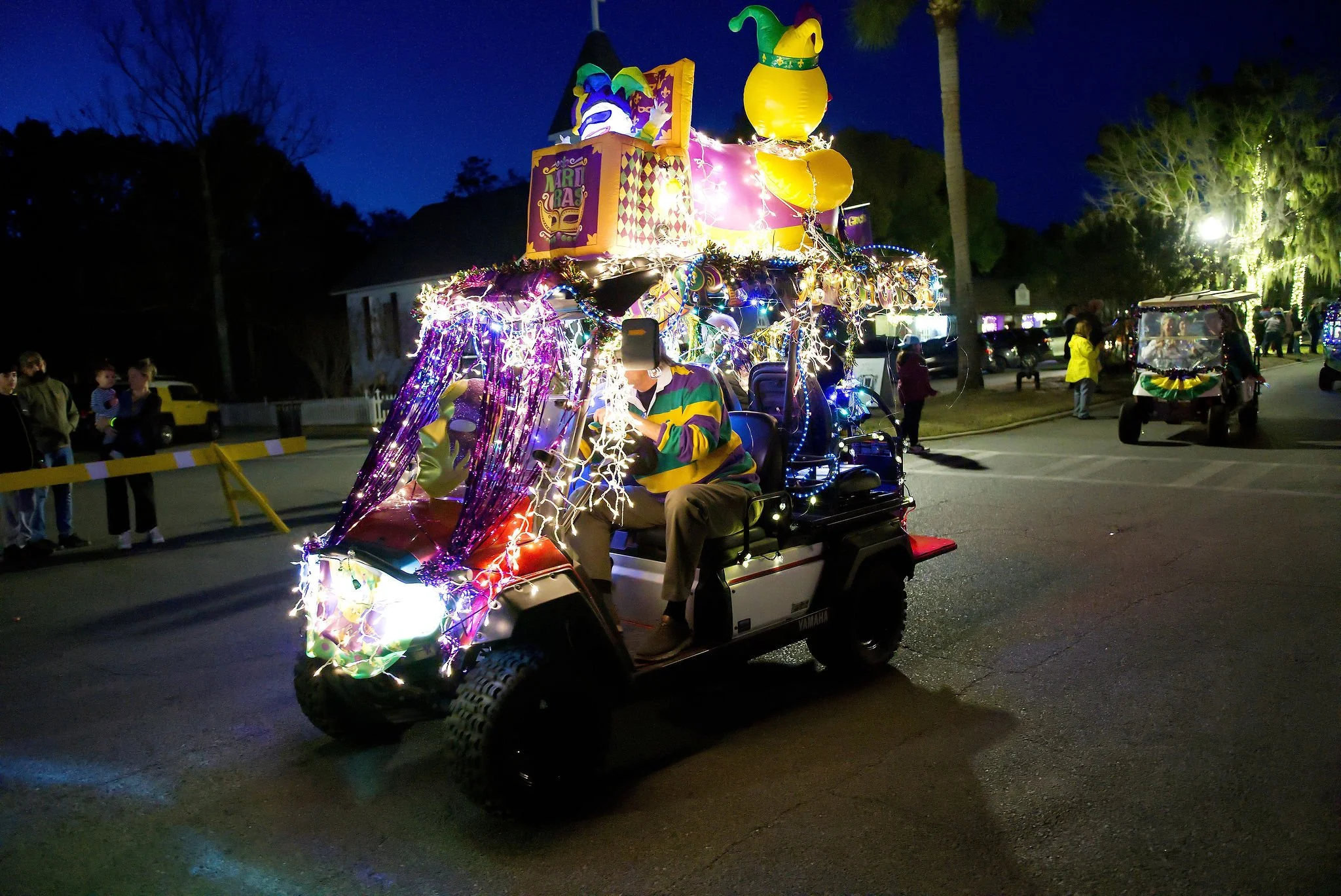 Golf cart decorated with colorful lights and Mardi Gras themed decorations, including a giant yellow mug with a green jester hat on top, during a parade at night.