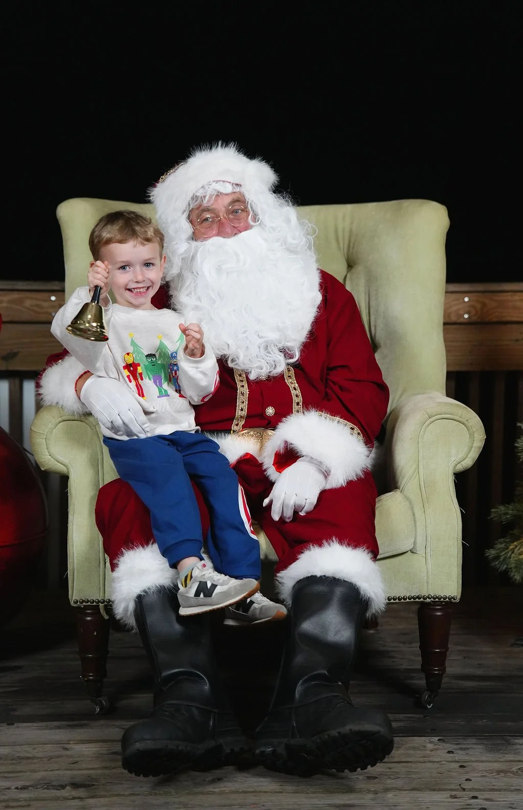 A young boy sitting on Santa Claus's lap, smiling, holding a bell. Santa is sitting in a green armchair, wearing a traditional red and white suit, with a large white beard and glasses. The background is dark, with a wooden floor and a Christmas tree 
