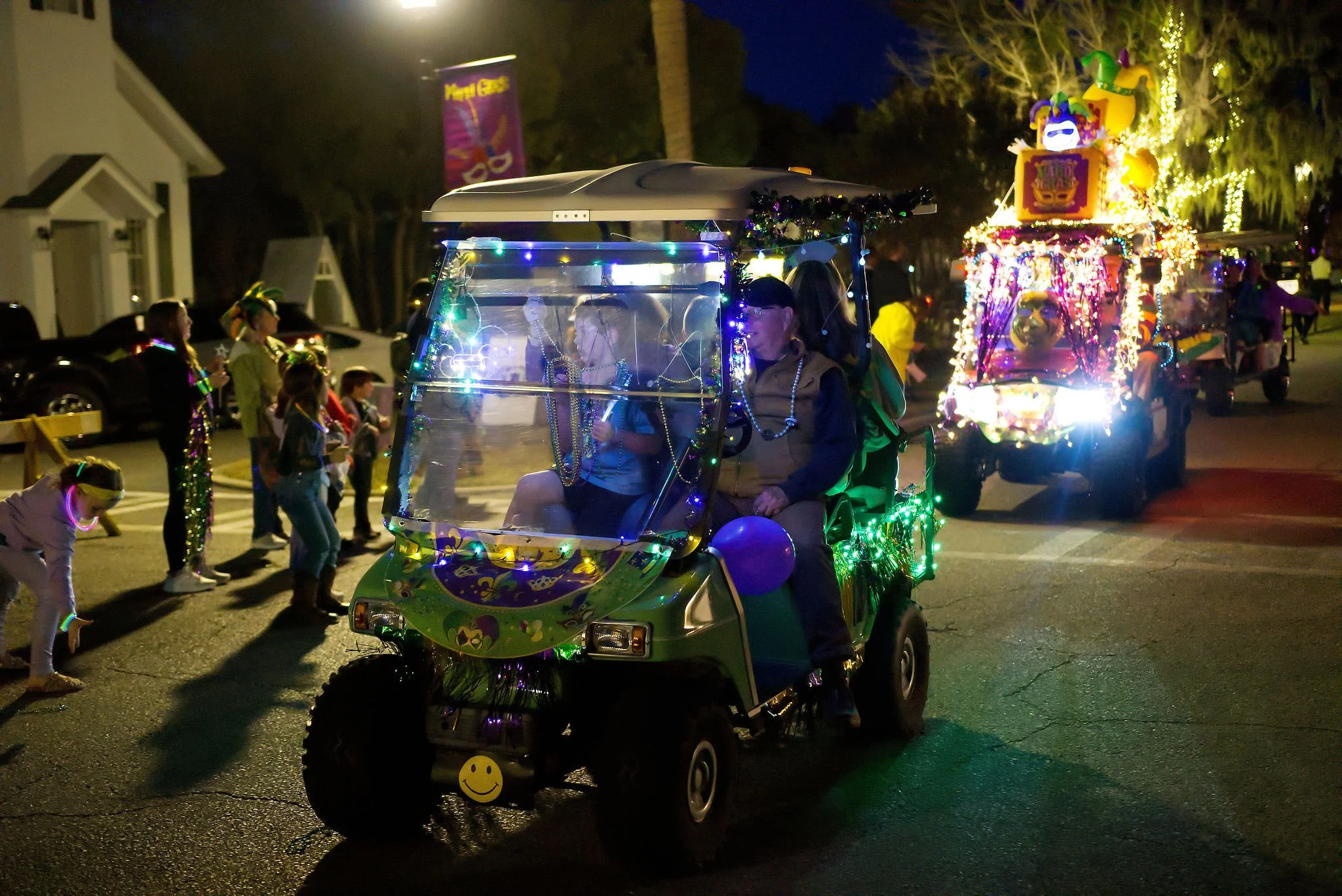 A decorated golf cart in a nighttime parade, illuminated with colorful lights, surrounded by children and spectators, with parade floats and people in the background.