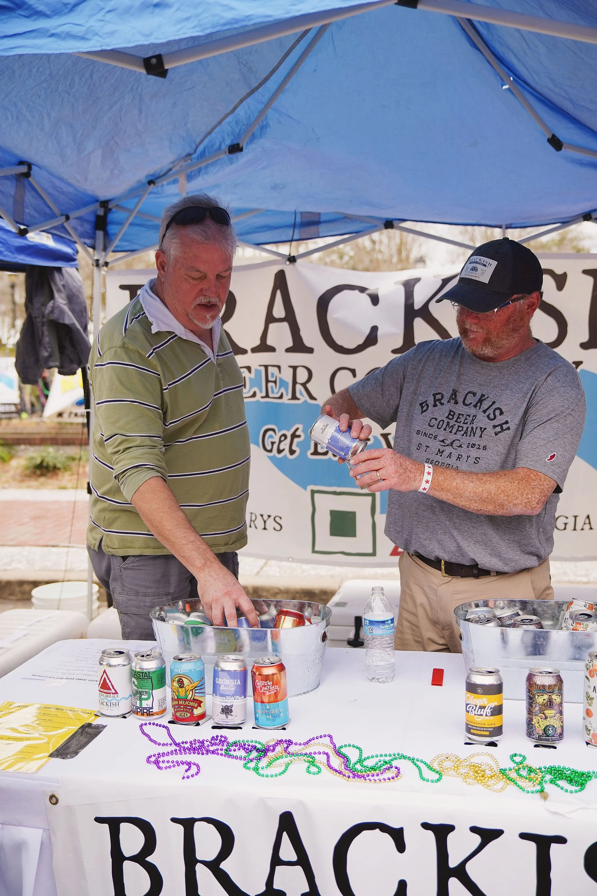 Two men under a blue canopy at a beer tasting event, with cans of craft beer and Mardi Gras beads on a table in front of them.