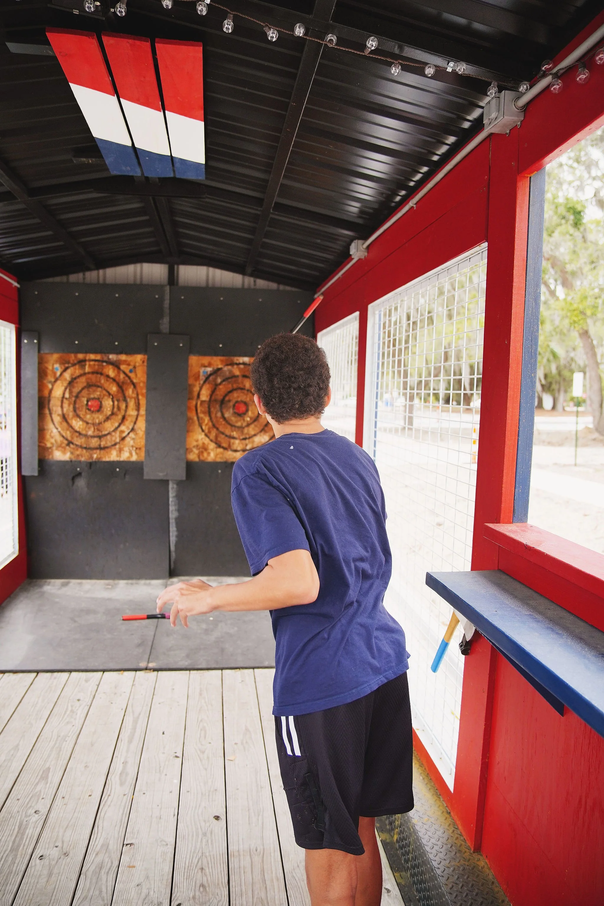 A young boy with curly hair wearing a dark blue t-shirt and black shorts is preparing to throw an axe at a target inside an enclosed axe-throwing booth. The booth has red and black walls, with a painted American flag on the ceiling. The background fe