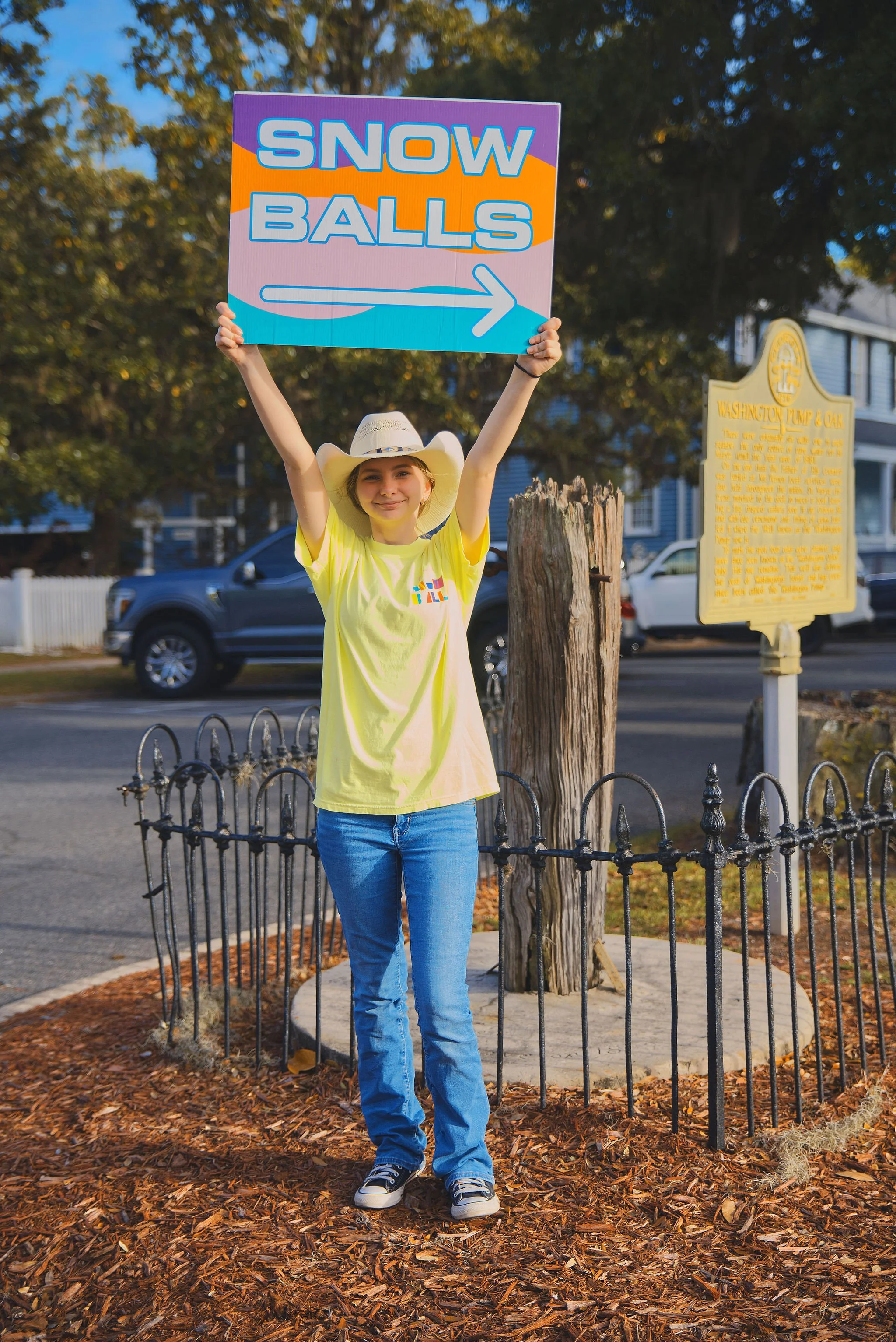 A young woman standing outdoors, wearing a yellow t-shirt and blue jeans, holding a colorful sign that says "Snow Balls" with an arrow pointing to the right. She is also wearing a white cowboy hat and smiling at the camera.