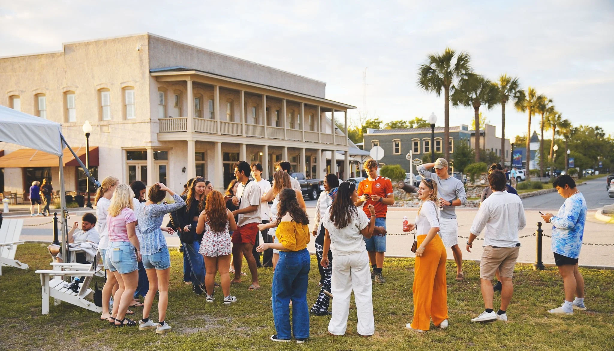 Group of people gathered outdoors on a grassy area in front of a white building with a balcony, palm trees, and parked cars, during sunset.