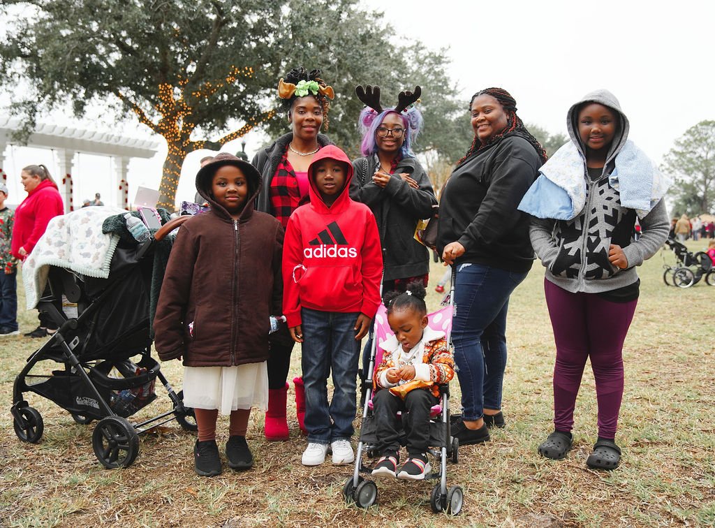 A group of seven girls and women outdoors at a park during daytime. One girl in a stroller wears a patterned jacket, and another girl is dressed in a brown hoodie and white skirt. The woman in the back center wears reindeer antlers and purple hair. T