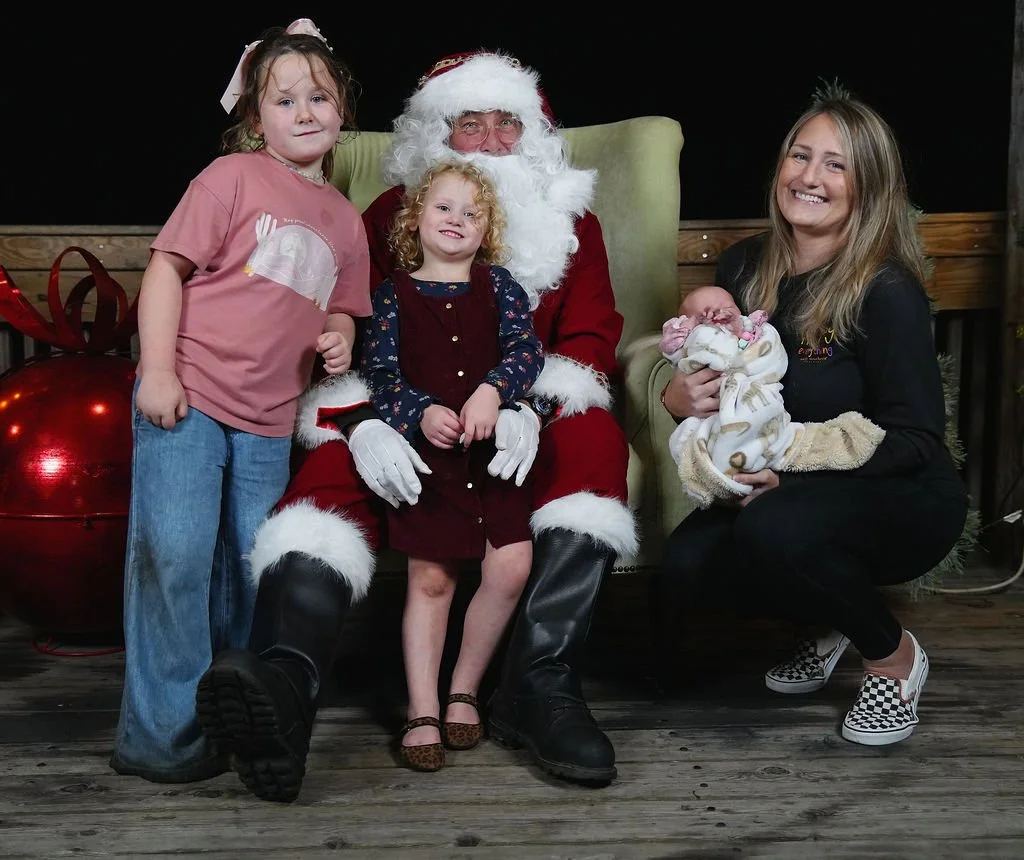 A Christmas photo with Santa Claus and children. Santa sits on a chair with two young girls, and a woman holding a newborn baby kneeling beside them. The background is dark with Christmas decorations.