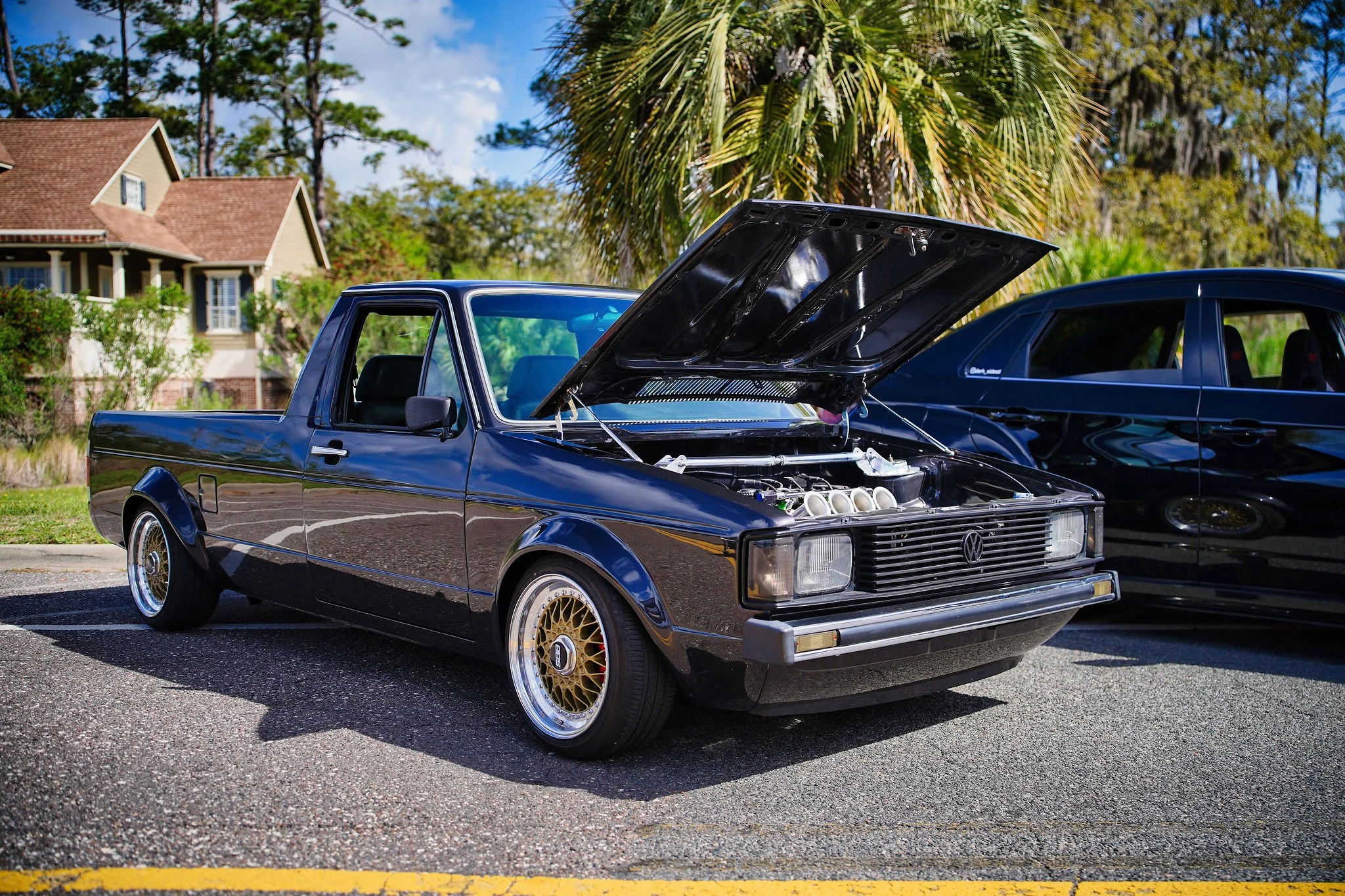 A classic blackVolkswagen car with its hood open, showing the engine, parked next to a modern black vehicle in an outdoor parking lot with a house, trees, and a palm tree in the background.