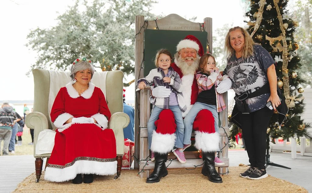 A group of people in Christmas costumes posing for a photo. Santa sits in the center with two children sitting on his lap. An older woman dressed as Mrs. Claus sits on a chair nearby. A woman in casual clothing stands next to Santa and the children, 