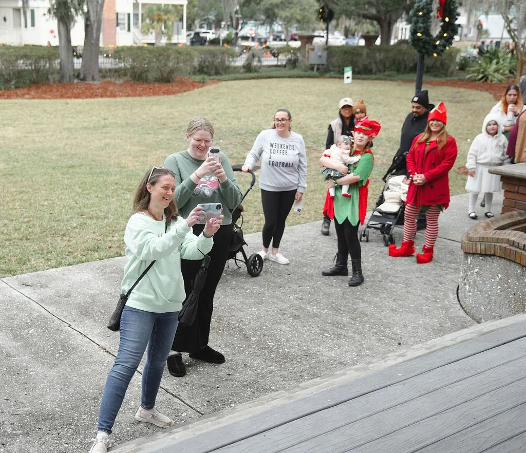 Group of people, some dressed in Christmas costumes, gathered outside in a park-like area, taking photos and smiling during a holiday gathering.