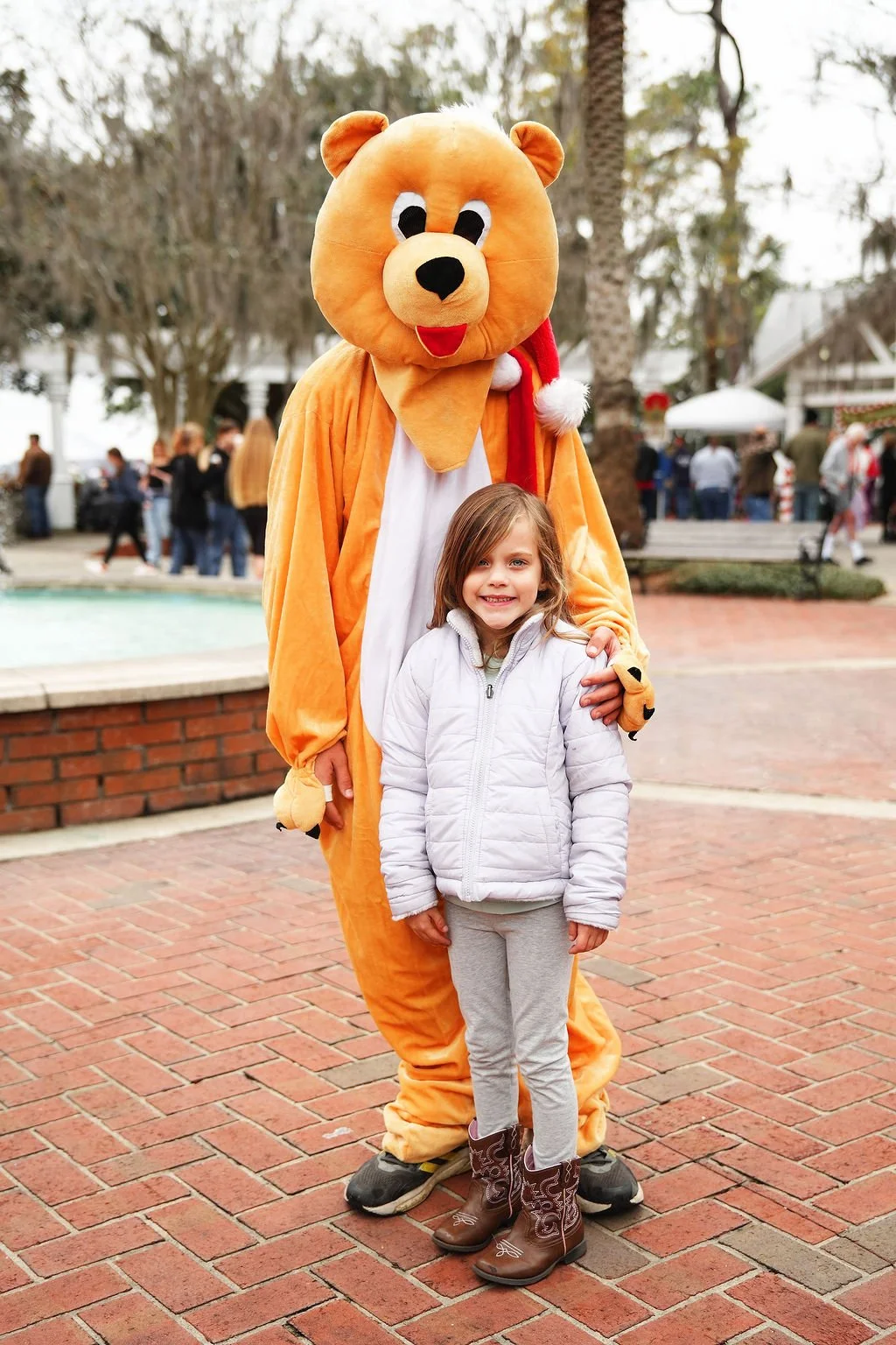 A young girl wearing a grey jacket and cowboy boots standing next to a person in a large bear costume with a Santa hat. They are outdoors on a brick-paved area surrounded by trees and a small crowd.