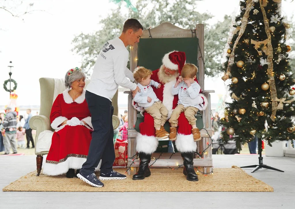 Children sitting on Santa's lap during a Christmas event, with a woman dressed as Mrs. Claus sitting nearby and a man assisting, all surrounded by holiday decorations including a Christmas tree.