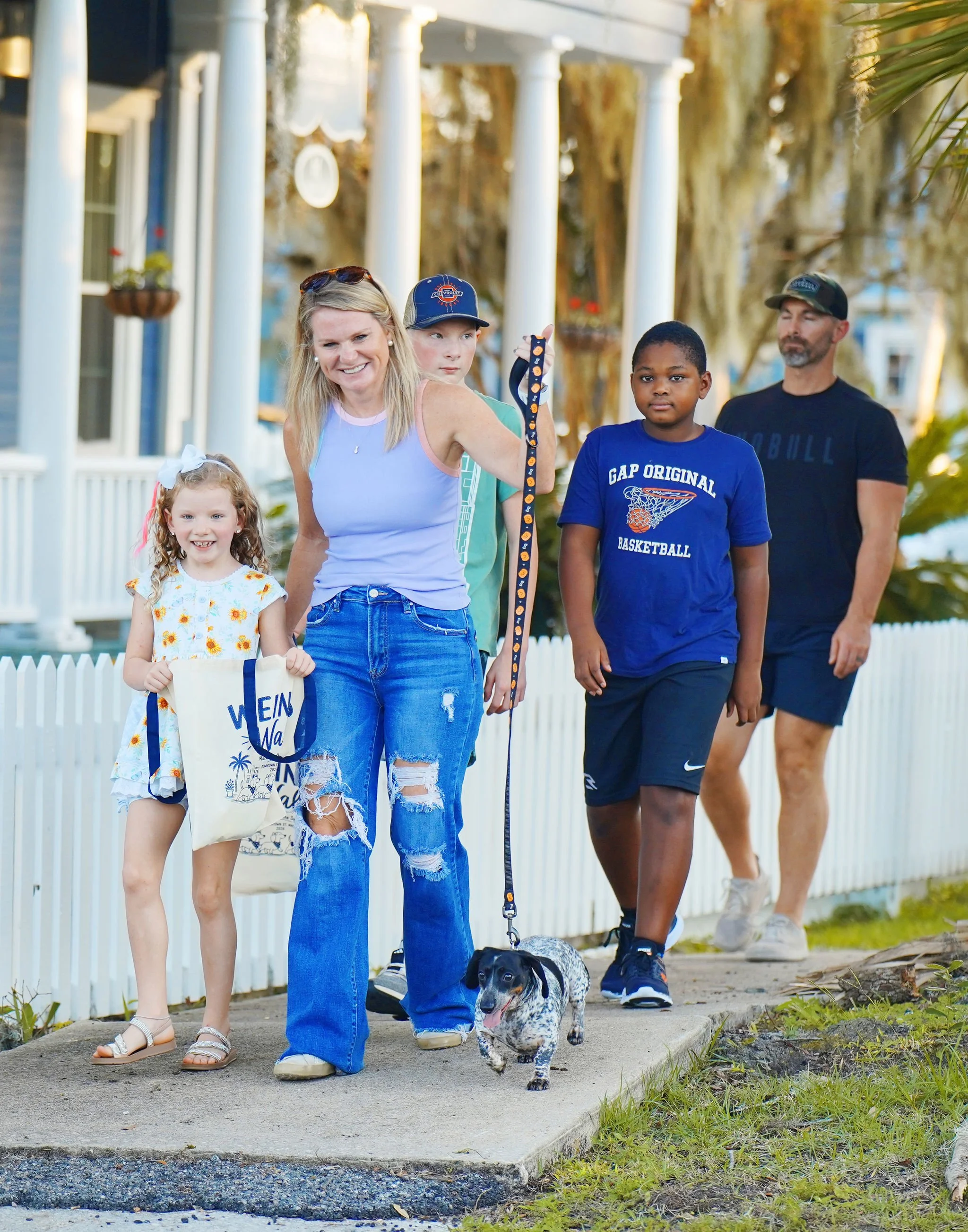 A group of people walking on a sidewalk, including two young girls, a woman, a teenage boy, and a man. The woman is holding a leash with a small dog, and the girls are smiling. The setting appears to be a residential neighborhood with white picket fe