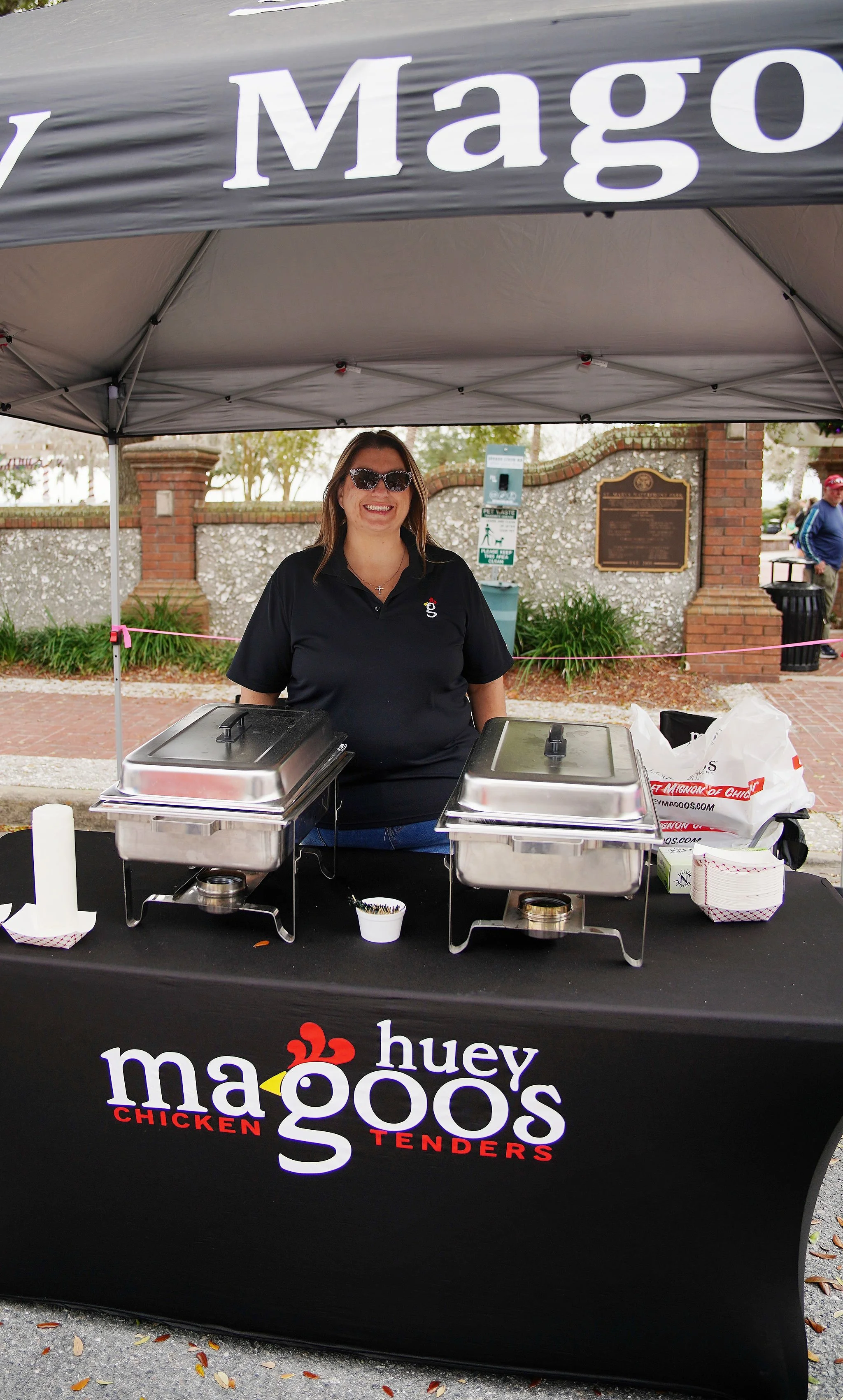 A woman standing behind a table at a food stand selling chicken tenders. She is smiling, wearing sunglasses and a black polo shirt with the logo for 'Huey Magoo's.' The tablecloth displays the restaurant's name and logo.