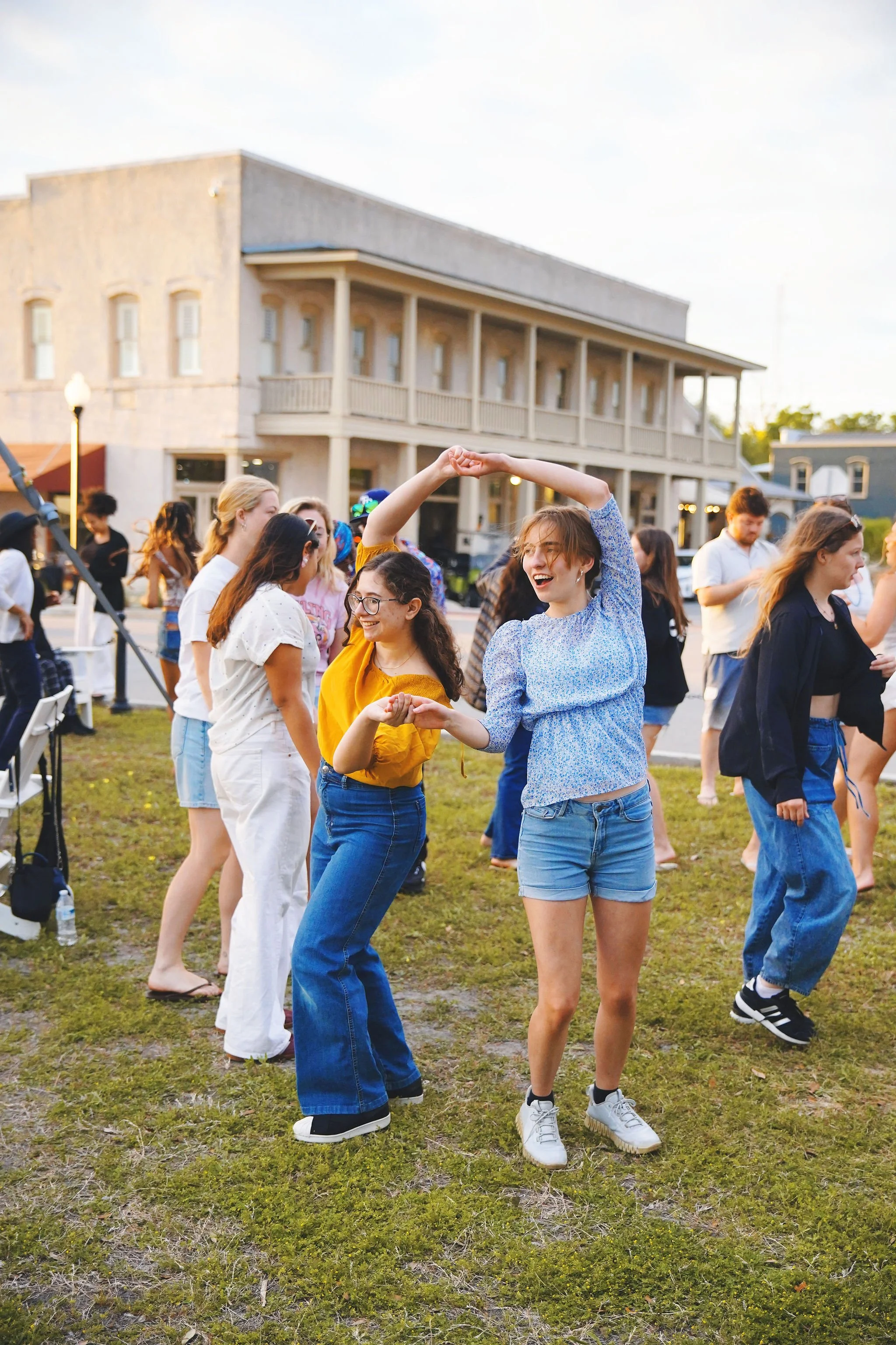 People dancing and having fun outdoors during sunset in a grassy area with buildings in the background.