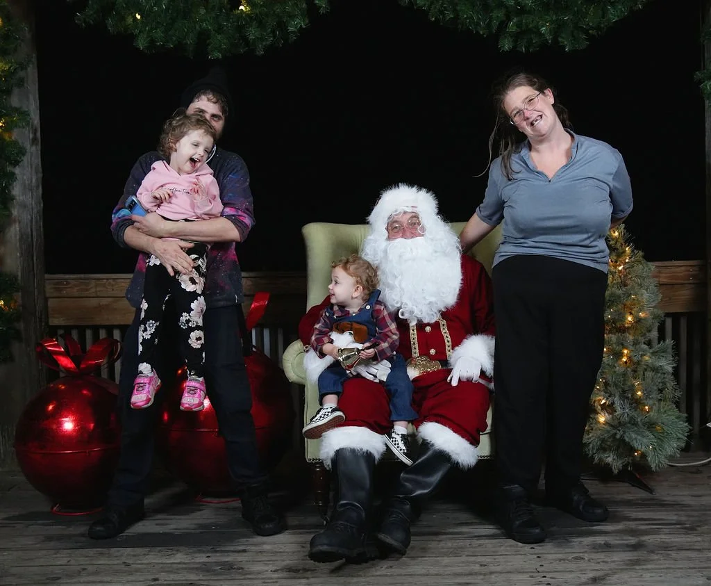 Family photo with Santa Claus, two children, and two adults, in front of Christmas decorations including a tree and large ornaments.