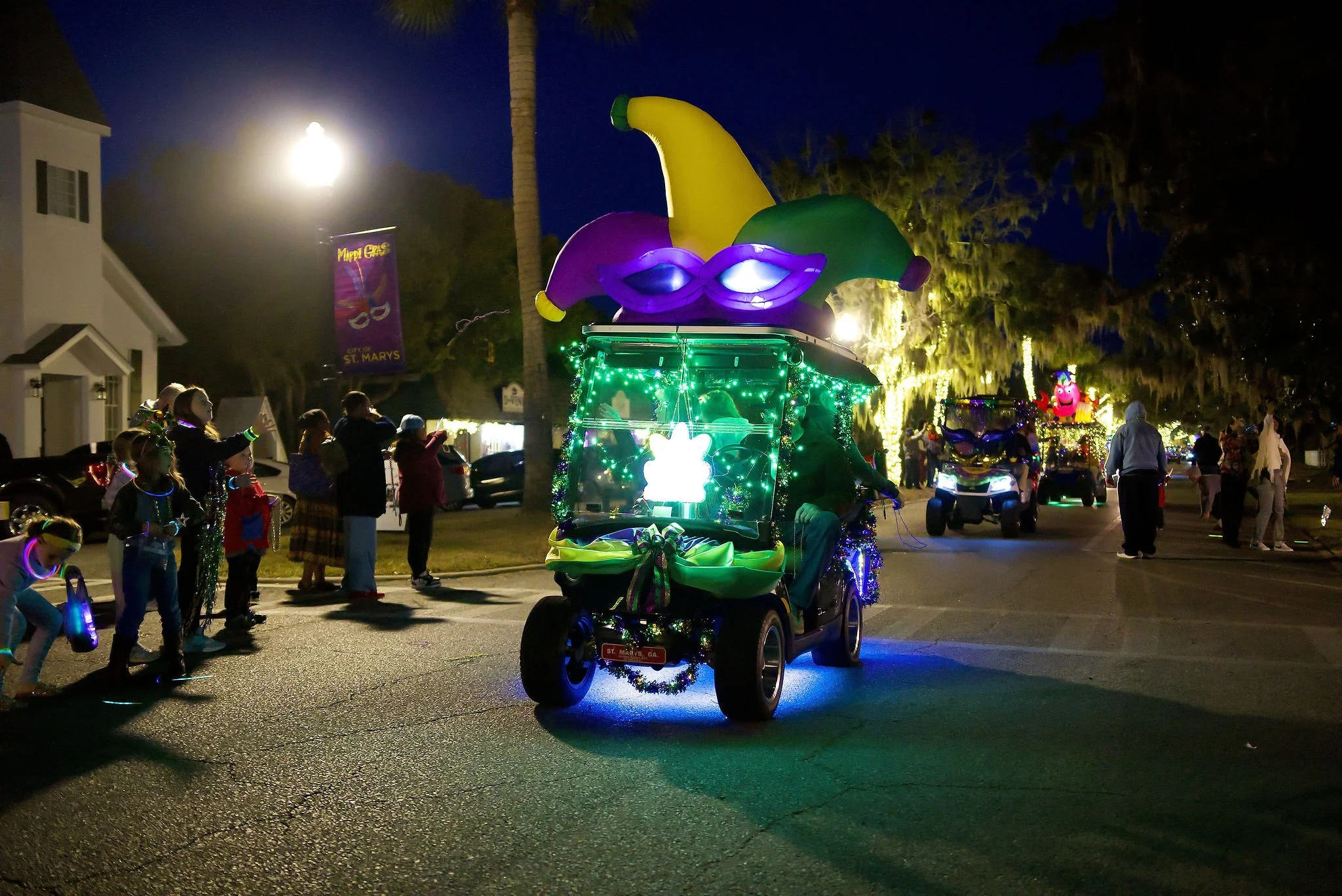 Nighttime parade with decorated golf carts, one in front with Mardi Gras-themed purple, green, and yellow decorations and large jester hat on top, followed by other decorated carts. Spectators stand along the street, some wearing glow necklaces, watc
