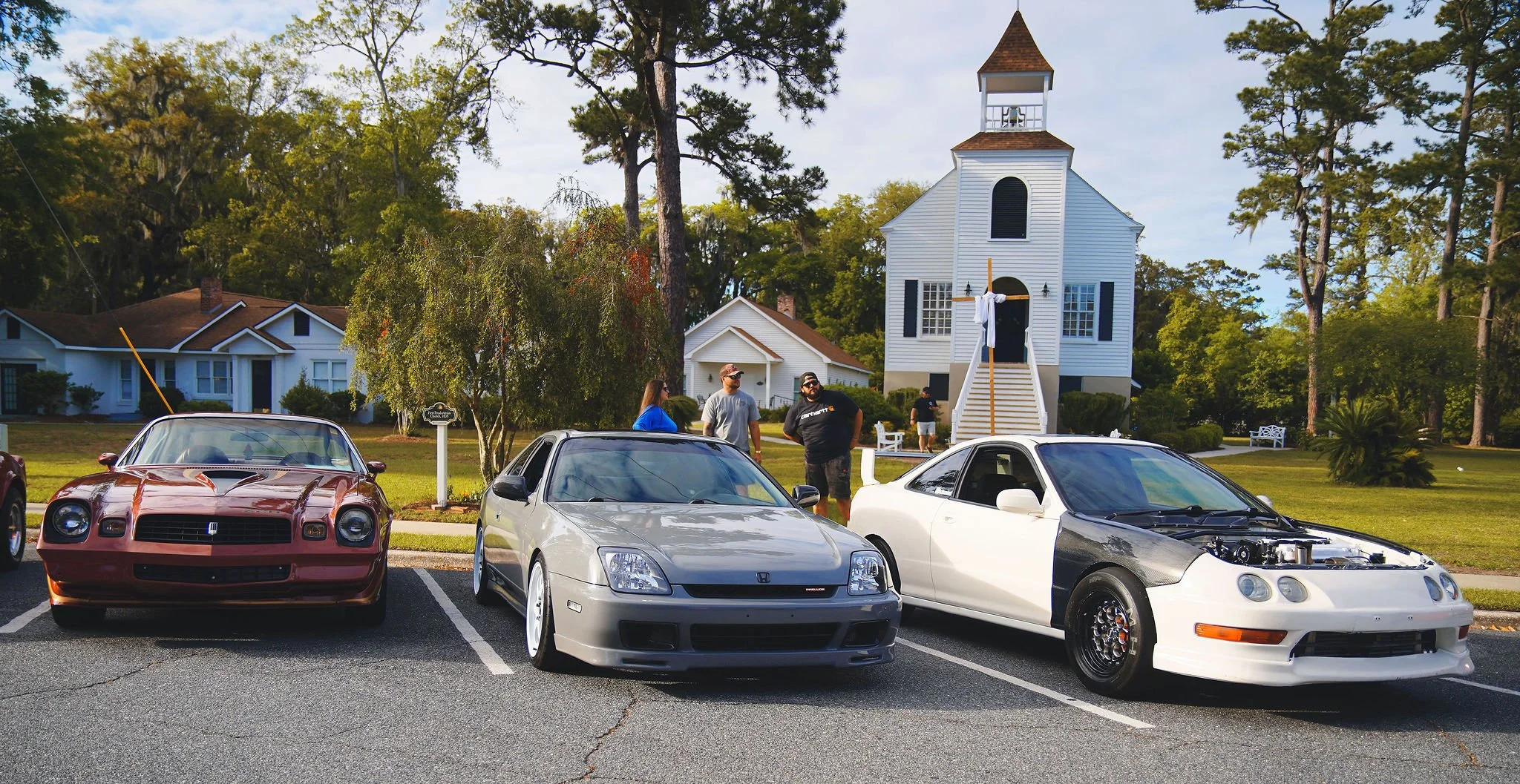 Three vintage cars parked in front of a white church with a steeple and a cross, with people standing nearby in a grassy area surrounded by trees and houses.