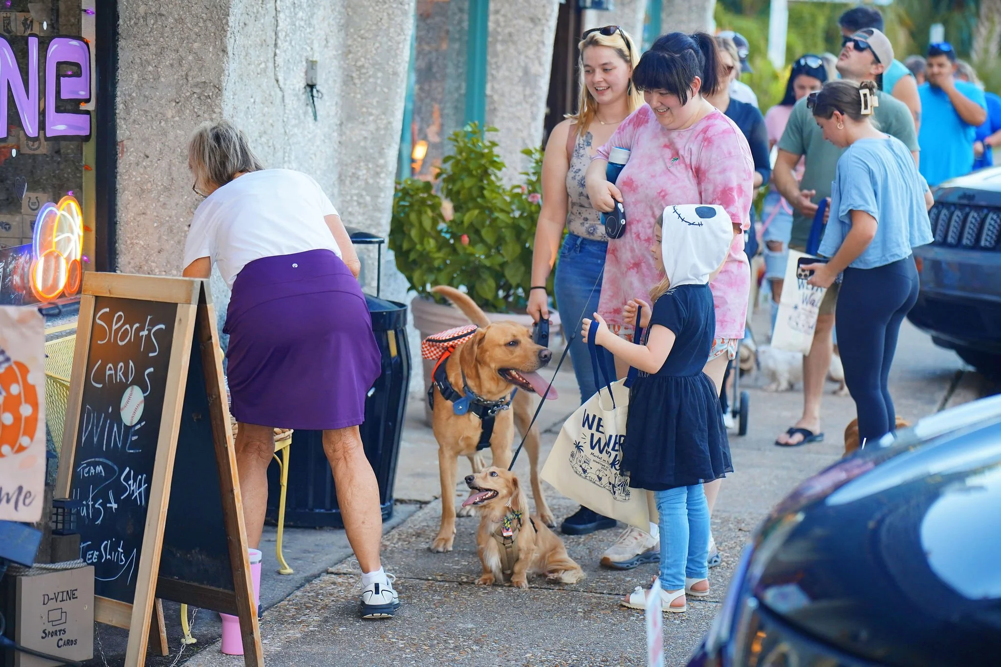 People lined up at an outdoor market or fair, talking and walking with dogs, including a young girl, women, and a man in the background. Some are wearing sunglasses and casual summer clothes. There are signs advertising sports cards and other items.