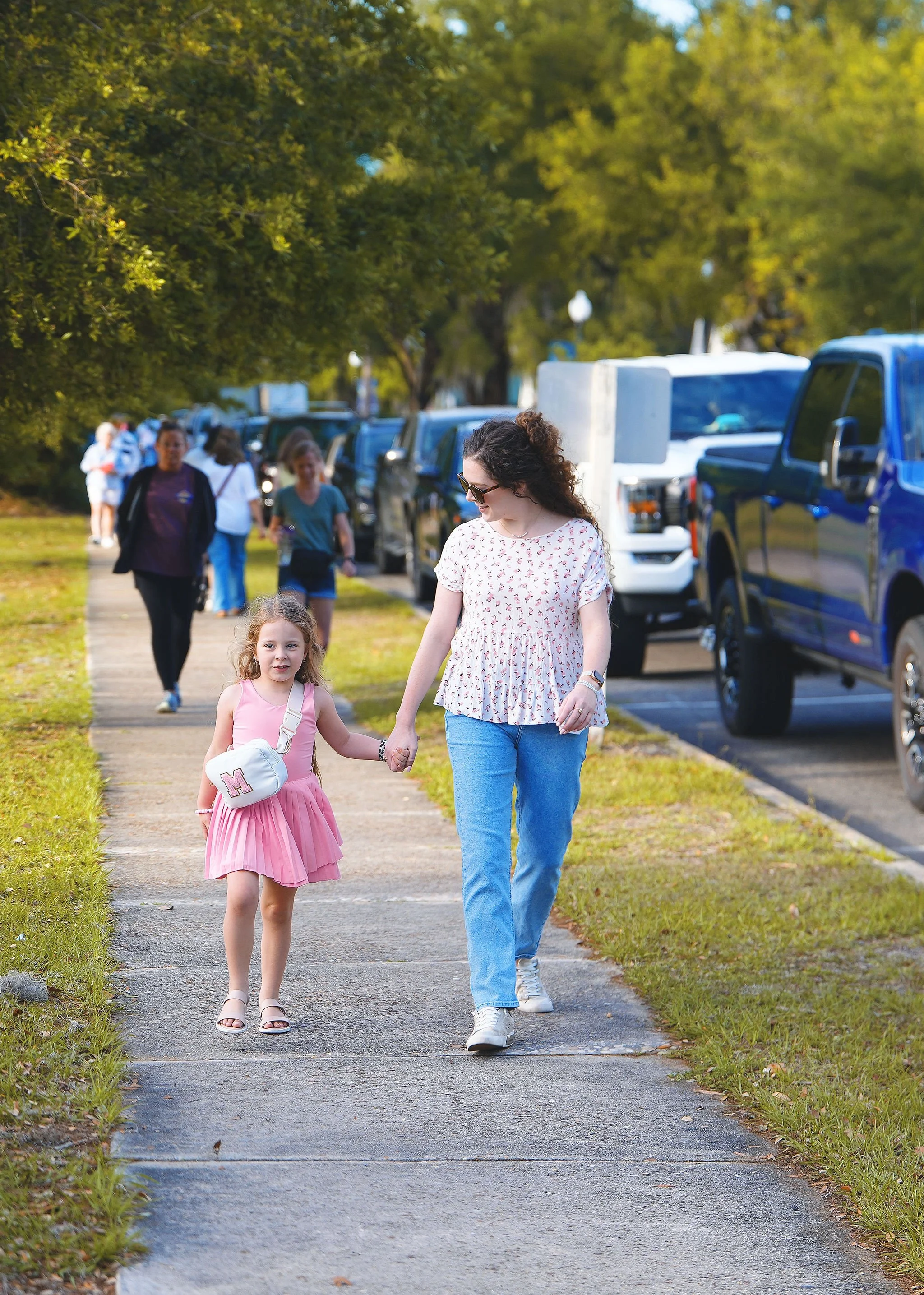 A woman and a young girl walk hand in hand on a sidewalk lined with parked cars on a sunny day, with trees in the background.