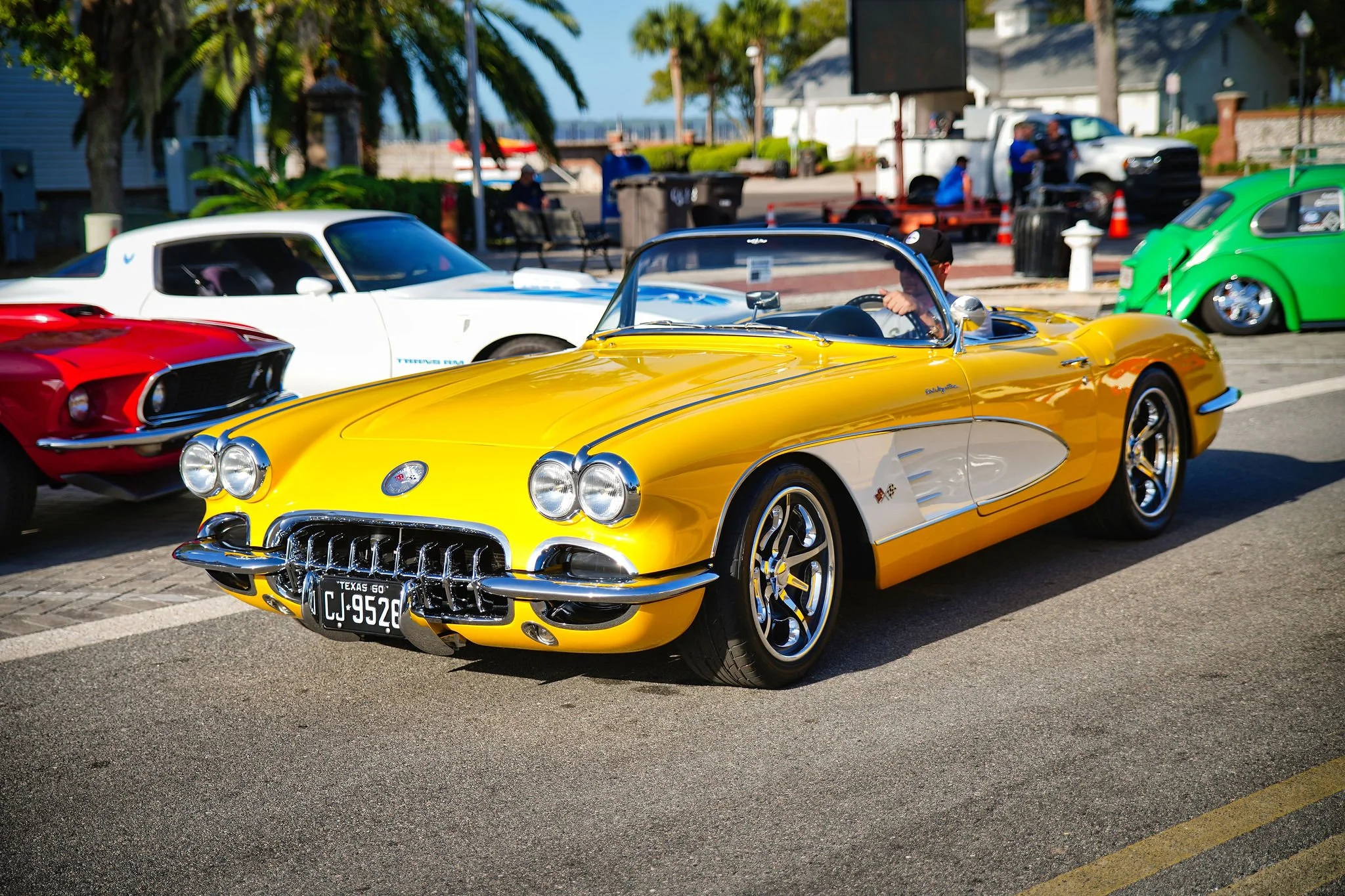 A yellow vintage Corvette convertible parked at a car show with other classic cars in the background, including a white car and a red car, at an outdoor event on a sunny day.