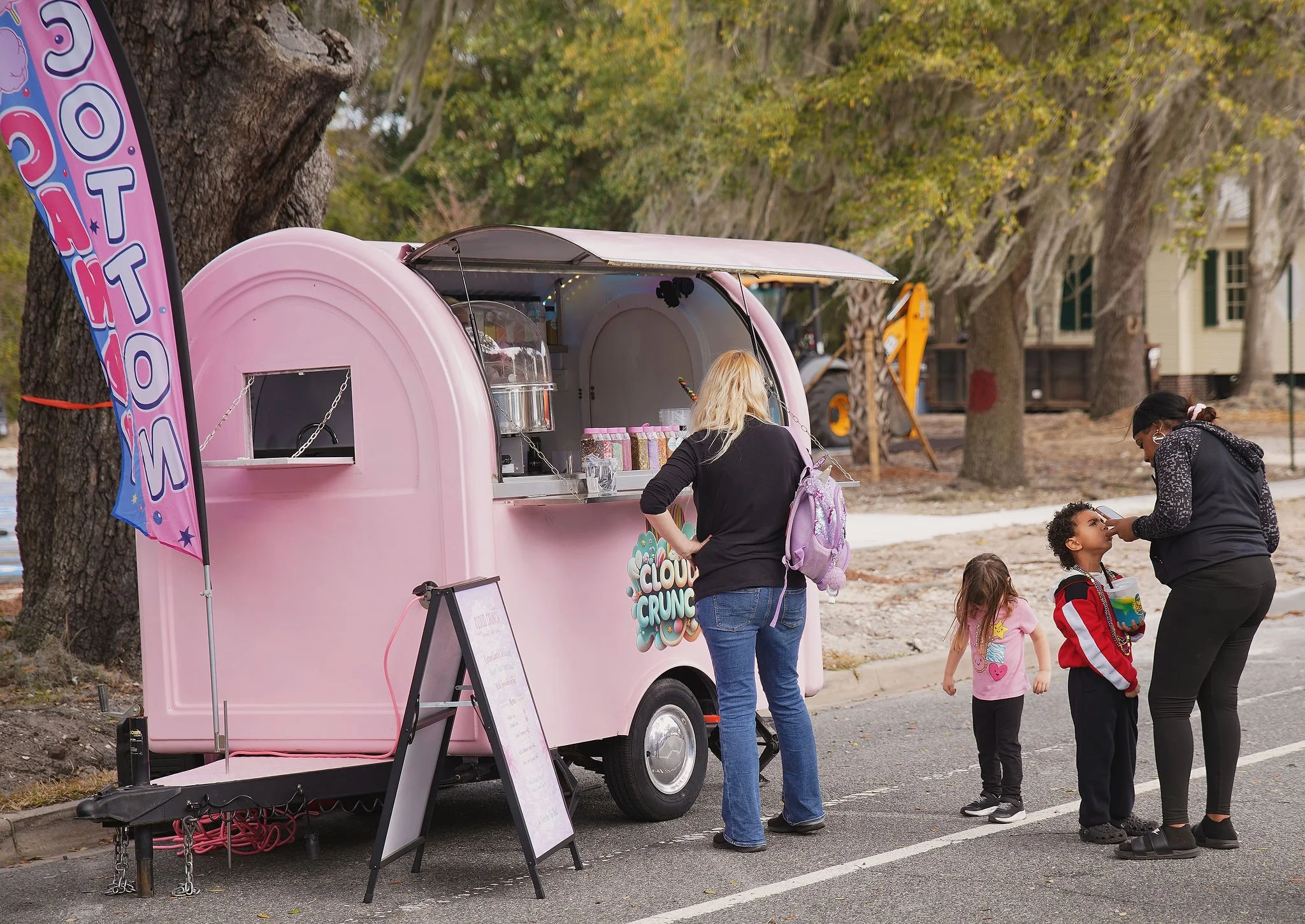 A pink cotton candy trailer labeled 'Cloud Crunch' stands on a street beside a large tree. A blonde woman in a black shirt and jeans is ordering or paying. Three children, including a girl in a pink shirt and two boys, are in line with a woman who is