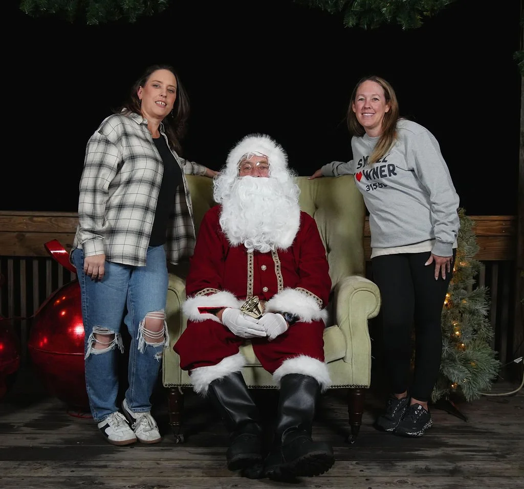 Two women standing beside Santa Claus, who is seated in a green armchair. The woman on the left wears a plaid shirt and ripped jeans, while the woman on the right wears a gray sweatshirt. Santa is dressed in a traditional red and white suit with a lo