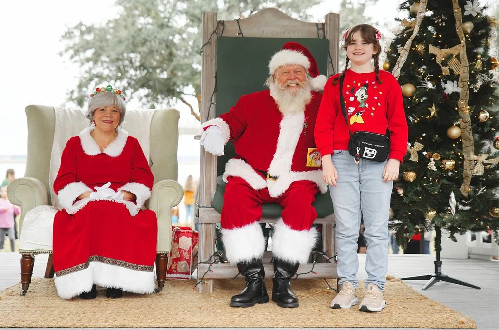 A girl standing next to Santa Claus and Mrs. Claus during Christmas, with a decorated Christmas tree in the background.