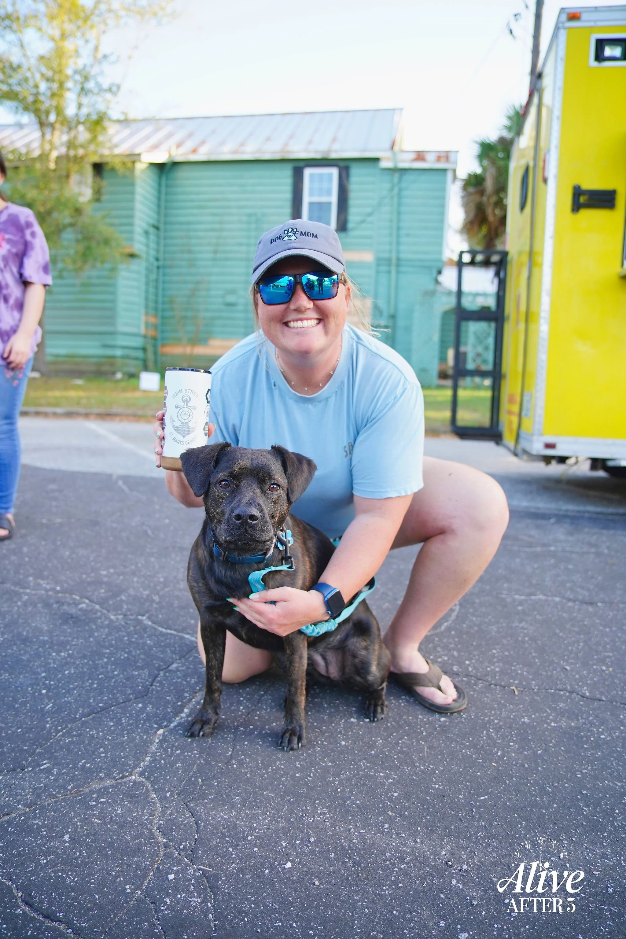 A woman squatting next to a black and brown dog on a cracked asphalt surface outside. She is smiling, wearing sunglasses, a gray hat, a light blue t-shirt, and flip-flops, holding a beverage. In the background, there is a green building, a yellow tra
