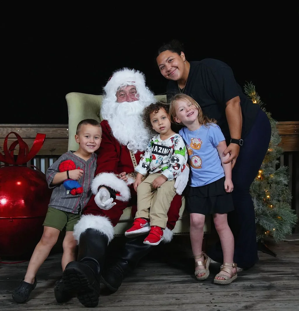 Children sitting on Santa Claus's lap with a woman standing next to them, a Christmas tree and large red gift box in the background.
