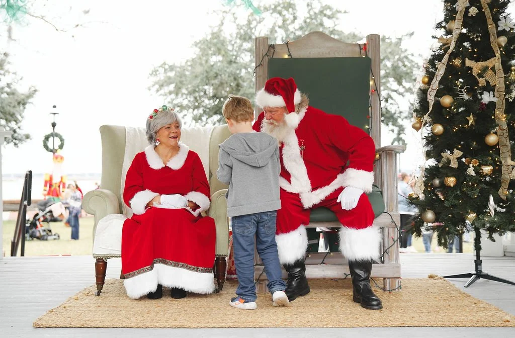 A young boy standing in front of Santa Claus and Mrs. Claus during a Christmas event outdoors. Santa is sitting on a chair, and Mrs. Claus is sitting on a vintage armchair. There is a decorated Christmas tree to the right and a chalkboard behind Sant