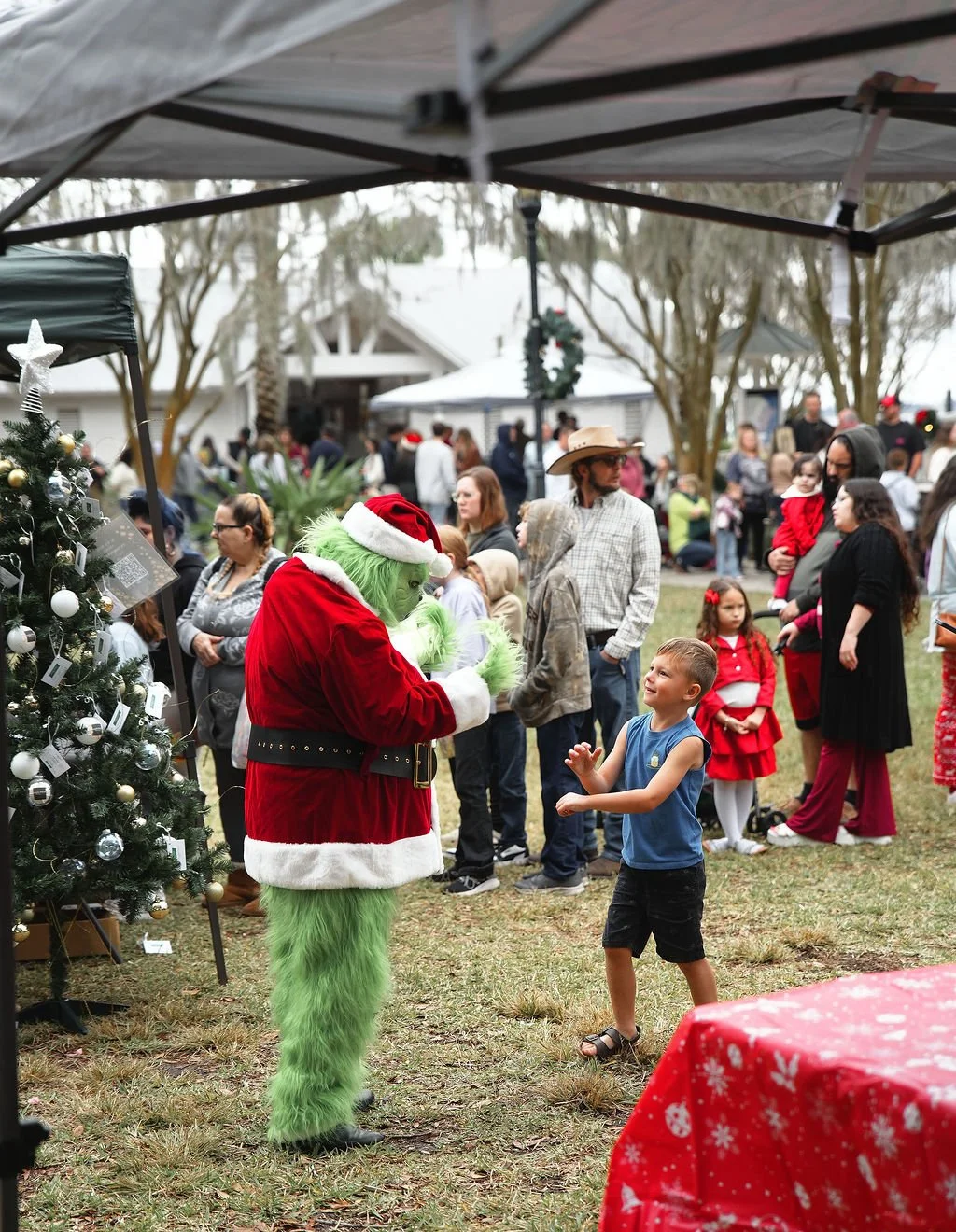 A young boy happily interacts with a person dressed as the Grinch in a Santa costume at a Christmas event outdoors, with a decorated Christmas tree nearby and many people gathered in the background.